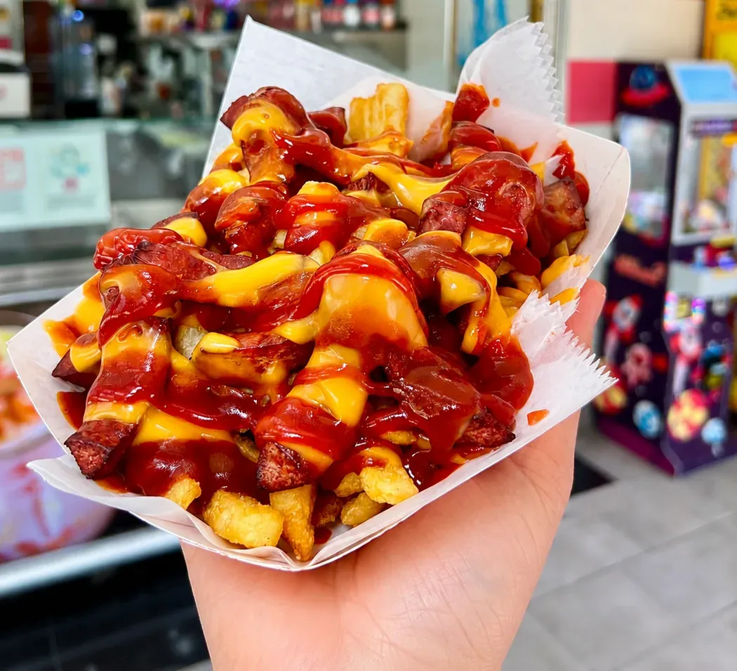 A paper tray of fries loaded with melted cheese sauce, chopped meat and red sauce, held up in front of the counter inside La Michoacana Premium Pilsen.