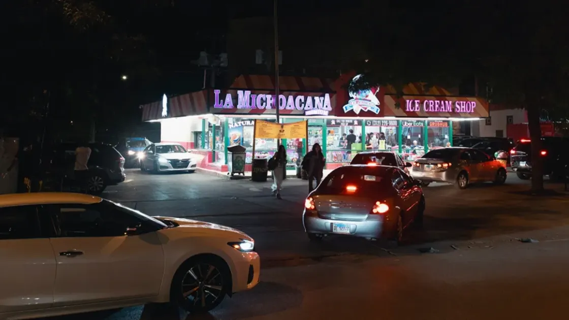 La Michoacana Premium Pilsen lit up after dark, with cars at the corner and people walking up to the counter windows.
