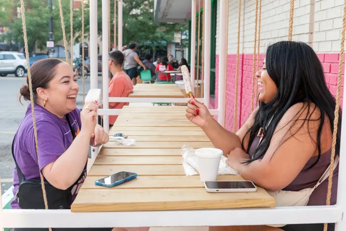 Two guests laughing over paletas at the swing seating outside La Michoacana Premium Pilsen.